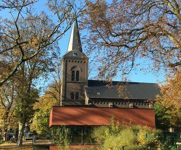 Eine Steinkirche mit hohem Turm, umgeben von Bäumen, mit einem neueren Gebäude davor.